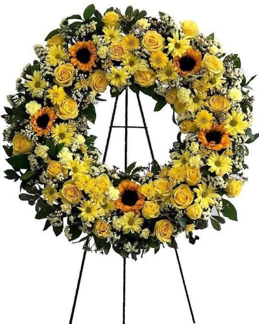 Funeral wreath arrangement with a large round design, adorned with vibrant sunflowers, yellow roses, mums, and carnations, complemented by accents of Regatta Piggie and Misty White. Enhanced with Rosal leaves and white statice foliage.