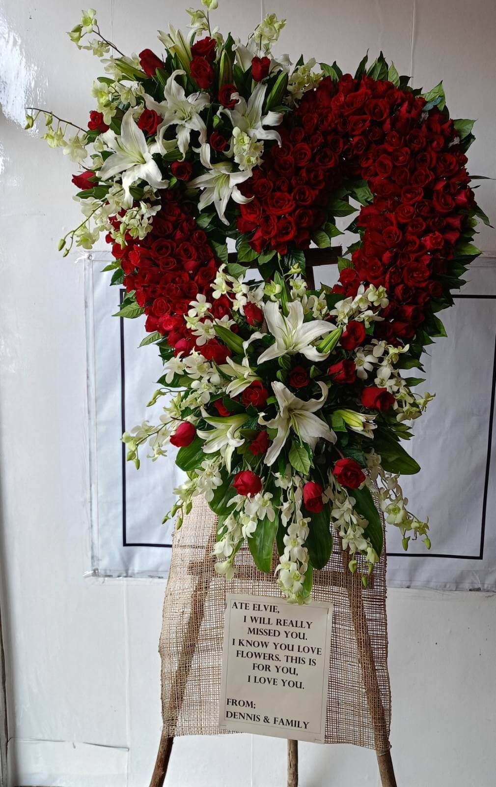 Heart-shaped flower stand for Purity & Spiritual Condolences. Featuring Red Roses, Casa Blanca lilies, and White Orchids flowers, accented with Rosal leaves, Greenline leaves, and Mathiola foliage, this elegant arrangement expresses heartfelt sympathy.